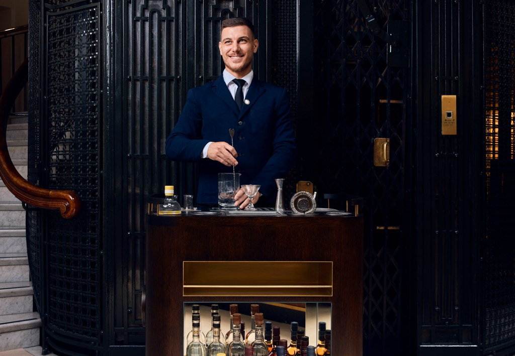 The image shows a man in a suit smiling behind a bar counter stocked with bottles of alcohol.